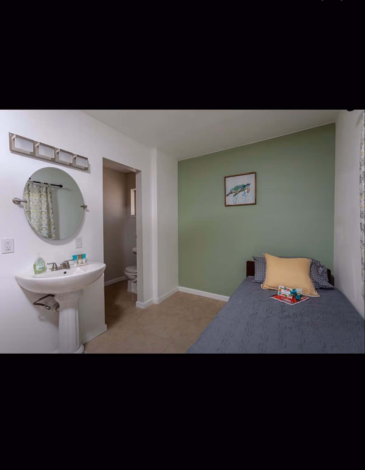 Small bedroom with a single bed against a green accent wall, a pedestal sink on the left and a doorway leading to a toilet.