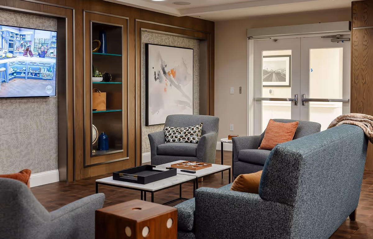 A cozy living room area with modern gray armchairs and a sofa, decorated with patterned and solid orange pillows. A wooden coffee table with a tray and a game board sits in the center. The room features a wall-mounted TV showing a cooking show, built-in shelves with decorative items, abstract wall art, and double doors with windows leading to another area.