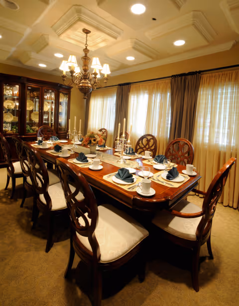 Formal dining room with a long wooden table set for dinner, upholstered chairs, chandelier, and a china cabinet.
