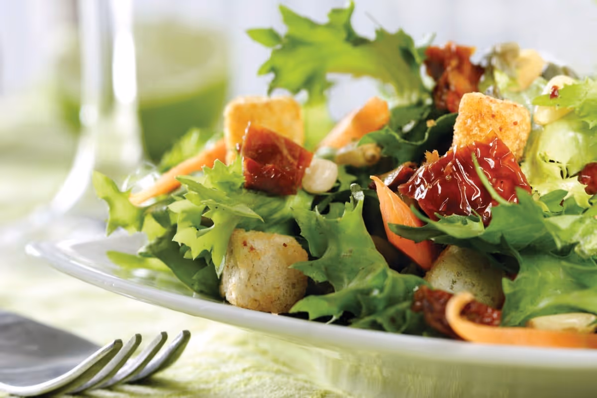 Close-up of a fresh green salad with leafy greens, croutons, sun-dried tomatoes, and carrot slices on a white plate with a fork nearby.