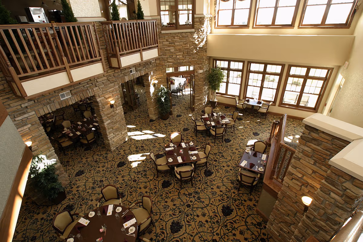 A spacious dining room with multiple round and square tables set with plates, cups, and napkins. The room features large windows allowing natural light to fill the space, stone pillars, a patterned carpet, and a wooden balcony railing overlooking the area.