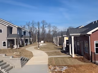 View of a residential senior living community with multiple single-story and two-story buildings along a curved concrete walkway, leafless trees in the background under a clear blue sky.