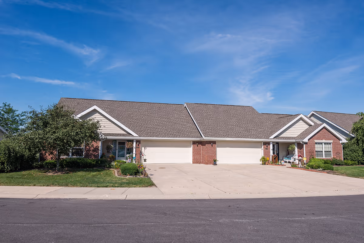 Front exterior of a single-story brick and siding senior living duplex with attached garages, driveways, and landscaped lawns under a blue sky.