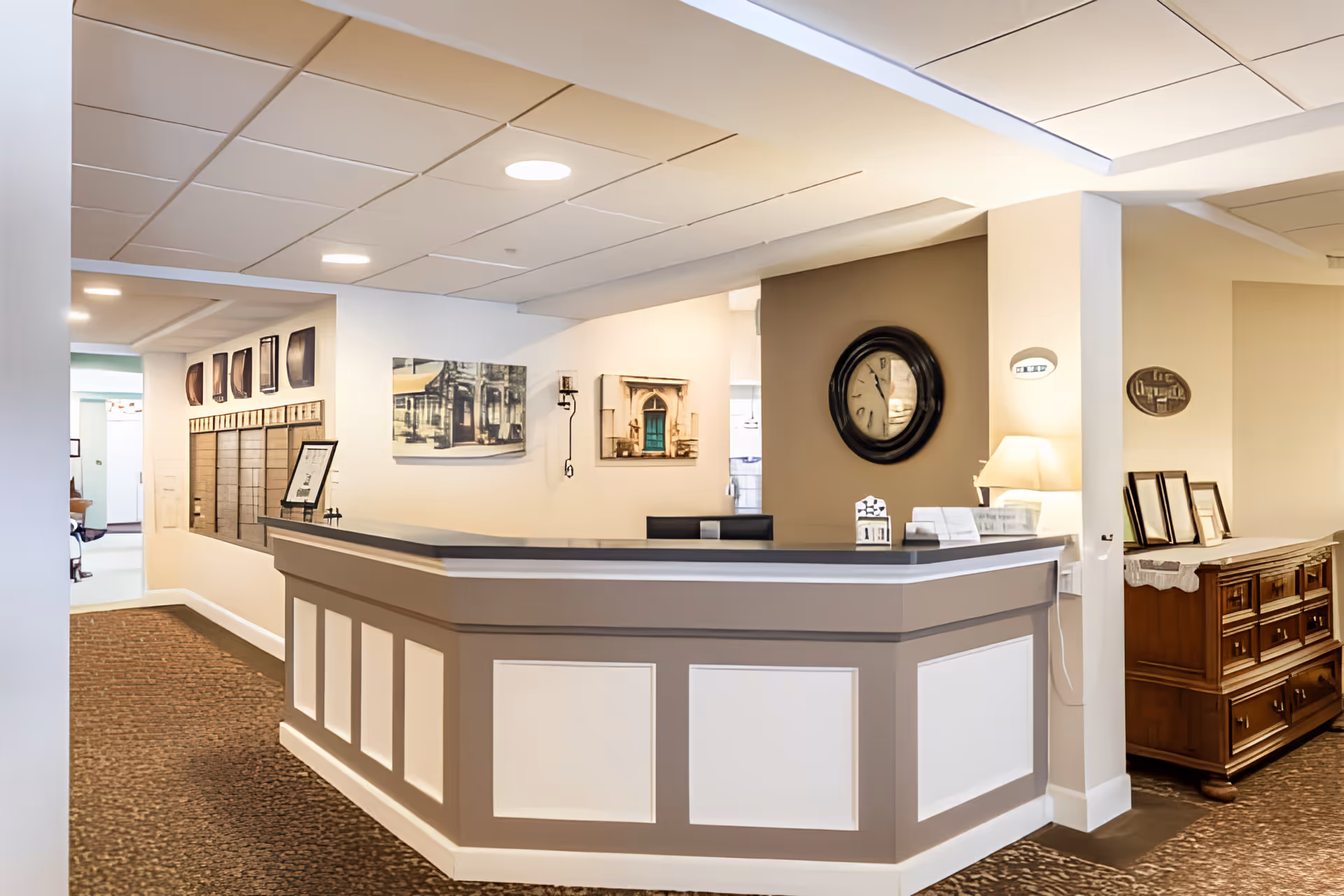 Reception desk and lobby area with a wall clock, artwork, and lamps in a senior living facility interior.