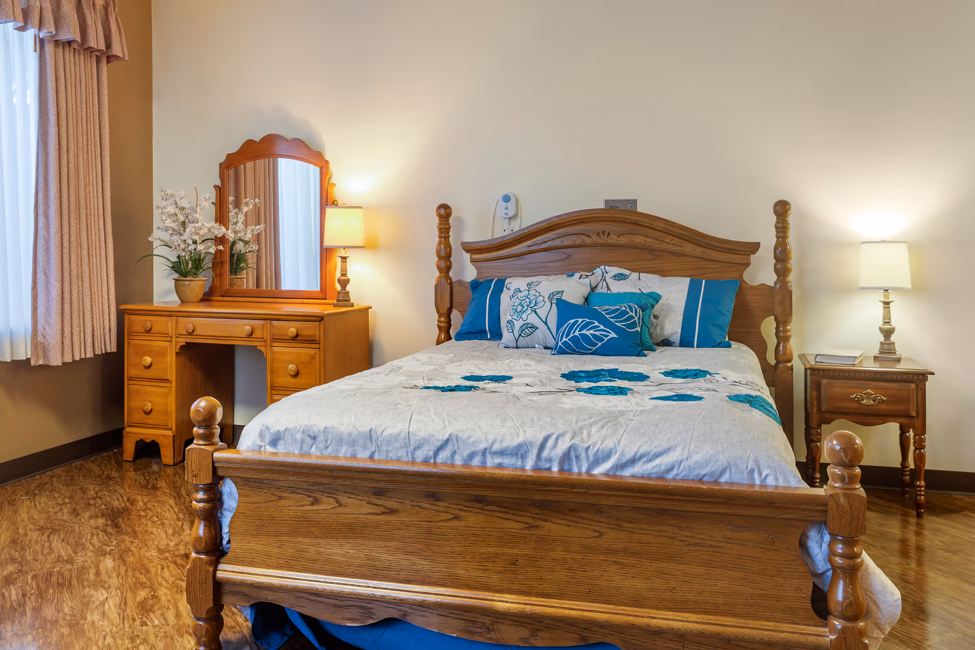Furnished bedroom with a wooden bed, matching nightstands and dresser with mirror, lamps, and floral bedding.