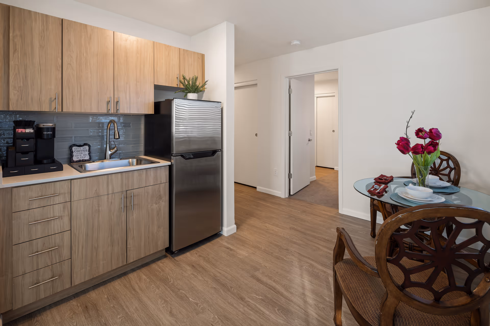 A small kitchen area with light wood cabinets, a stainless steel refrigerator, and a sink. There is a coffee maker and a small decorative sign on the counter. To the right, there is a round glass dining table with two wooden chairs and a vase with red flowers. The floor is wood, and there is a doorway leading to another room.