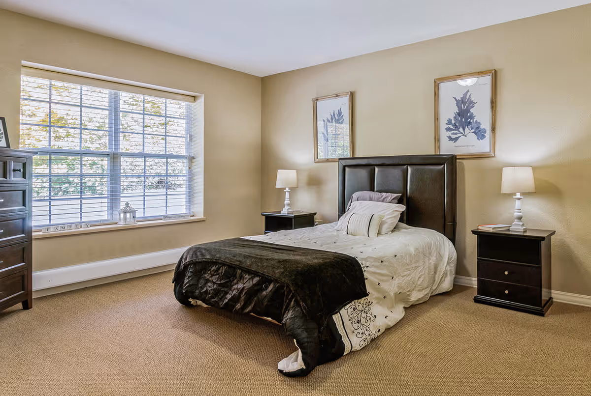 A cozy bedroom with a large window featuring white blinds, a bed with a dark headboard and white bedding with a black throw blanket, two matching nightstands each with a white lamp, and two framed botanical prints on the wall above the bed.
