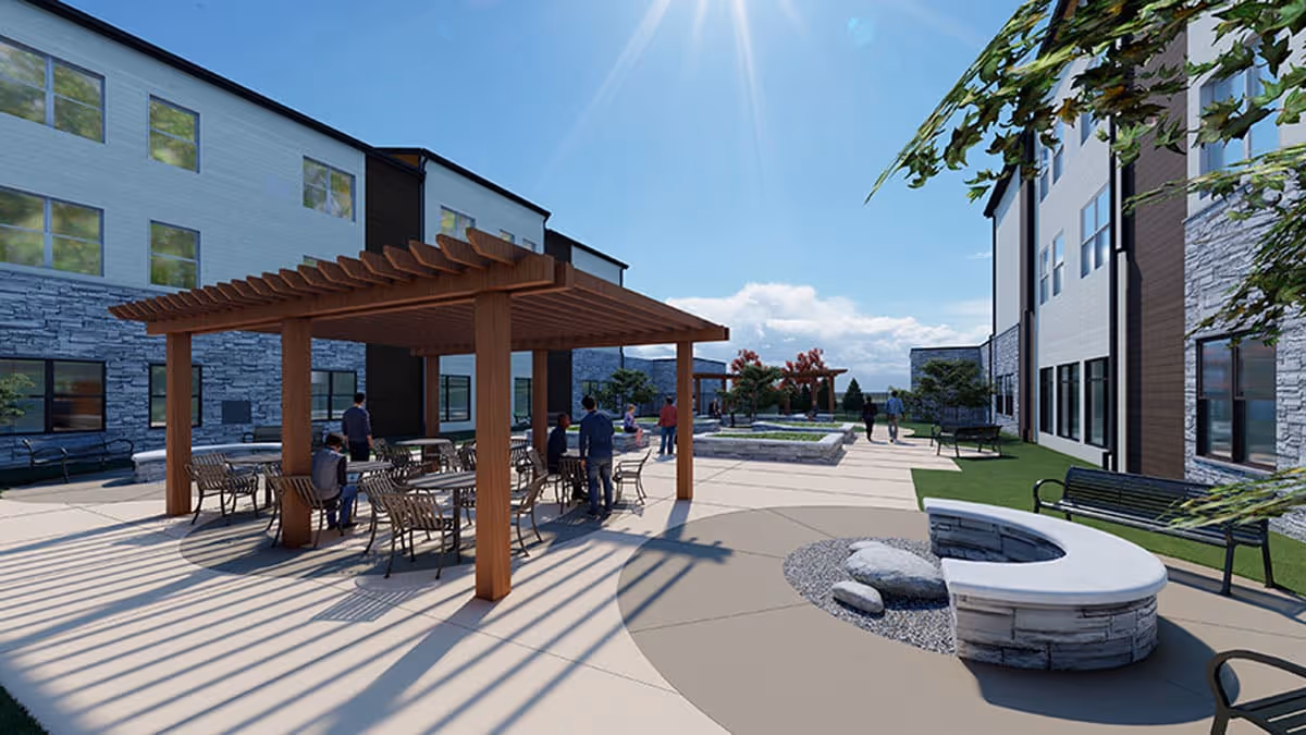 Outdoor patio area at a senior living facility with a wooden pergola covering tables and chairs. Several people are seated and standing around the area. The space includes benches, a curved stone fire pit, and landscaped greenery with a clear blue sky overhead.