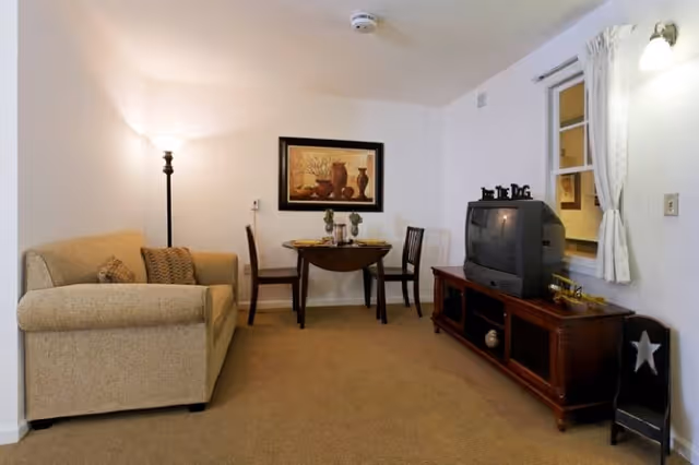 A cozy living room area featuring a beige sofa with two patterned cushions, a small wooden dining table set with two chairs and tableware, a wooden TV stand with an older-style CRT television, and a window with white curtains. The walls are white, and there is a floor lamp and a framed artwork of pottery on the wall.