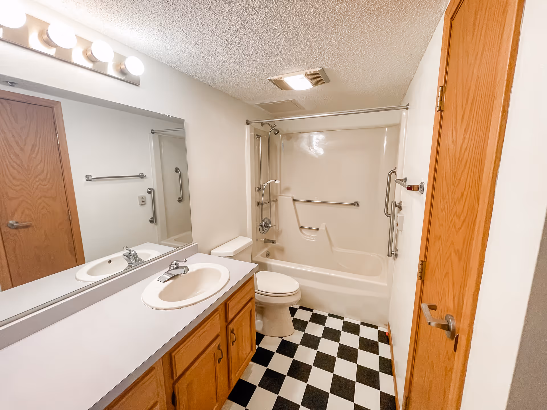 A bathroom with a white sink and countertop, wooden cabinets underneath, a large mirror above the sink with four round lights, a toilet, and a bathtub with a built-in seat and grab bars. The floor has a black and white checkered pattern, and there is a wooden door on the right side.
