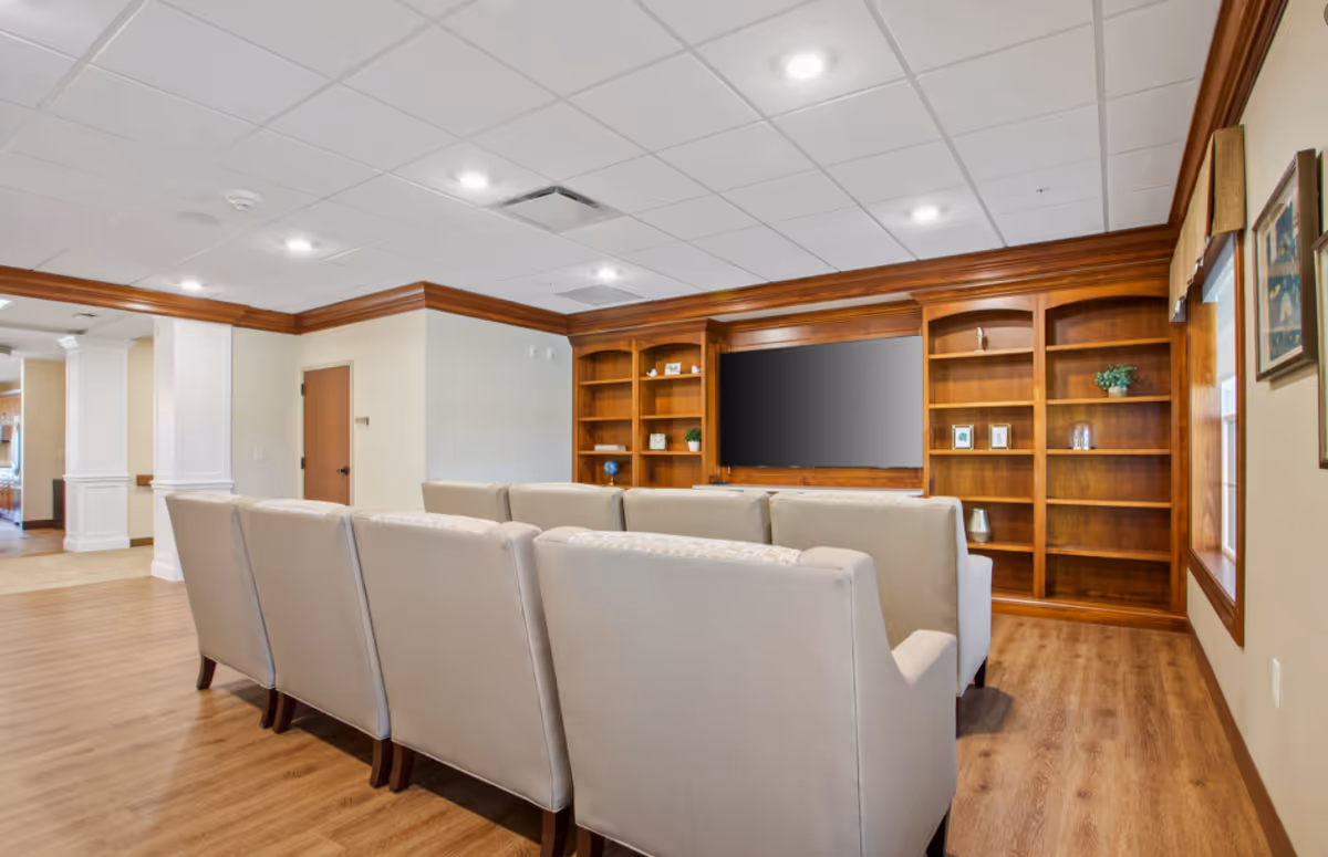 A communal seating area with rows of beige upholstered chairs facing a wall-mounted TV set within wooden built-in shelving in a senior living lounge.