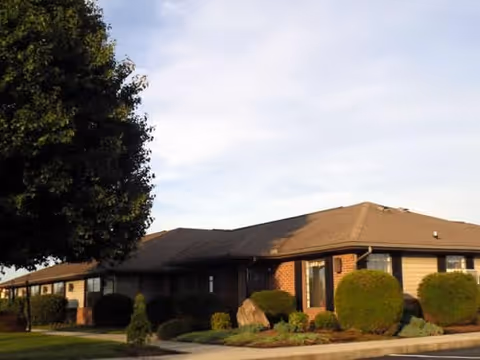 Single-story brick and siding building with a sloped roof, surrounded by neatly trimmed bushes and a tree, under a partly cloudy sky.