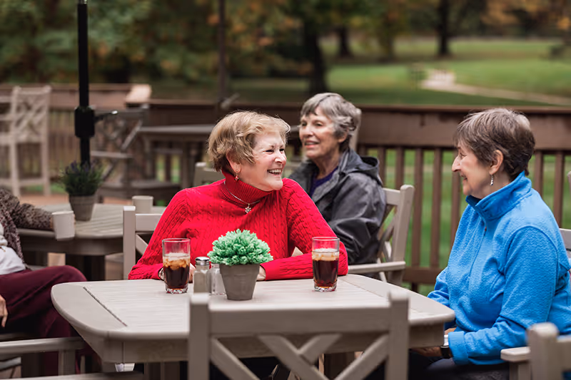 Three elderly women sitting at an outdoor wooden table on a patio, smiling and engaging in conversation. Two glasses of iced tea and a small potted plant are on the table. The background shows greenery and trees.