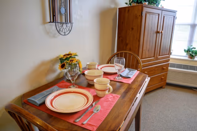 A small wooden dining table set for two with red placemats, plates, bowls, mugs, silverware, and glasses. A vase with sunflowers is on the table. In the background, there is a wooden cabinet and a window with blinds and a potted plant on the windowsill.