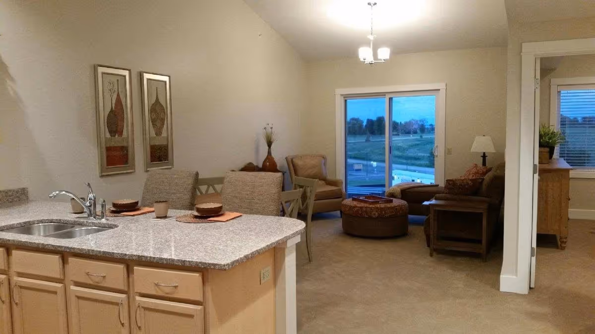Open-plan interior showing a kitchen island with sink and place settings, dining chairs, and a living area with armchairs and a sliding glass door.