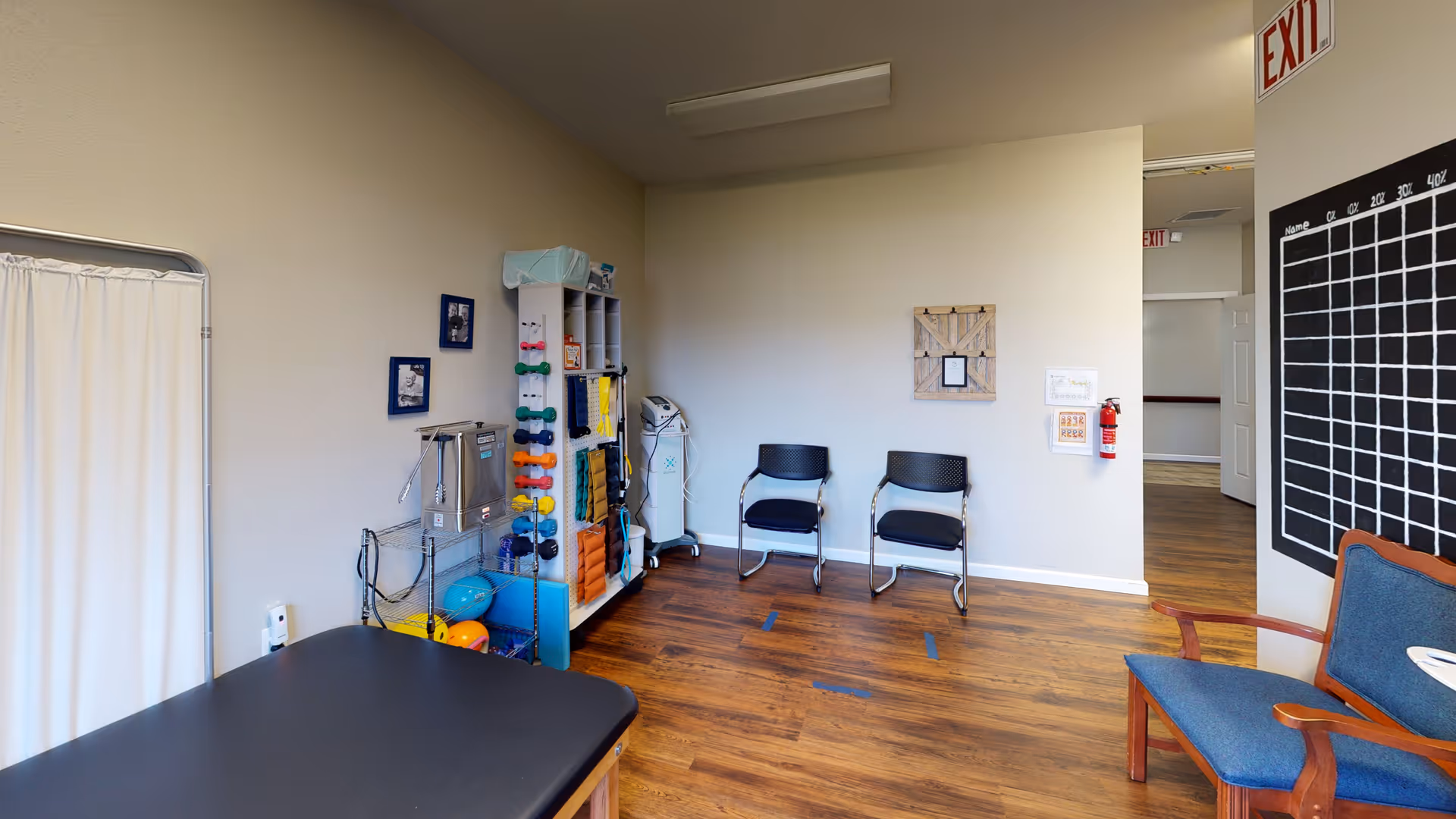 A therapy or exercise room with wooden flooring, two black chairs against a beige wall, a shelf holding colorful exercise equipment such as dumbbells, balls, and foam rollers, a black treatment table, and a blue cushioned chair. There is a white curtain on the left side and an exit sign above a doorway on the right.