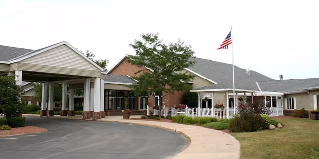Exterior view of North Terrace Assisted Living facility showing a single-story building with a covered entrance, a curved driveway, landscaped greenery, and an American flag on a flagpole.