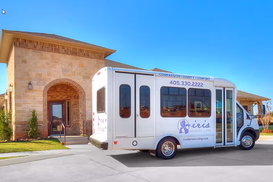A white shuttle bus parked in front of a stone and brick building under a clear blue sky. The bus has the logo and text for Iris Memory Care, including a phone number and website. The building has a large arched doorway and some small bushes near the entrance.