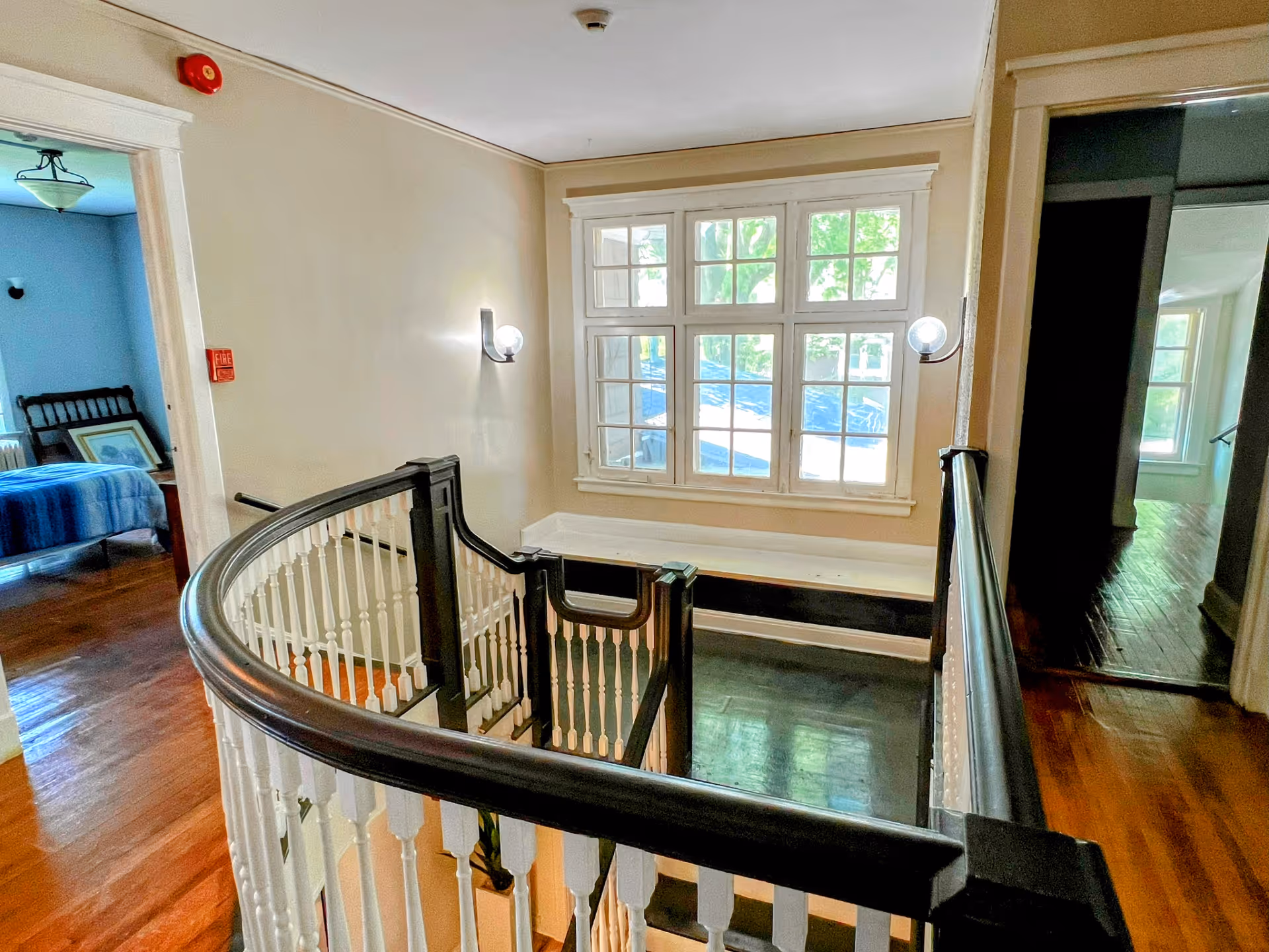 Interior view of a senior living facility showing a wooden staircase with black handrails and white balusters. There is a large window letting in natural light, and two wall-mounted lights on either side of the window. To the left, a bedroom with a bed covered in a blue blanket is visible. The floor is wooden throughout.