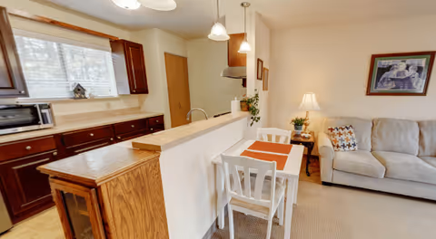 Interior view of a senior living facility showing a small dining area with a white table and two chairs, a beige sofa with patterned pillows, a side table with a lamp and plant, and a kitchen area with dark wood cabinets, a microwave, and a window with blinds.