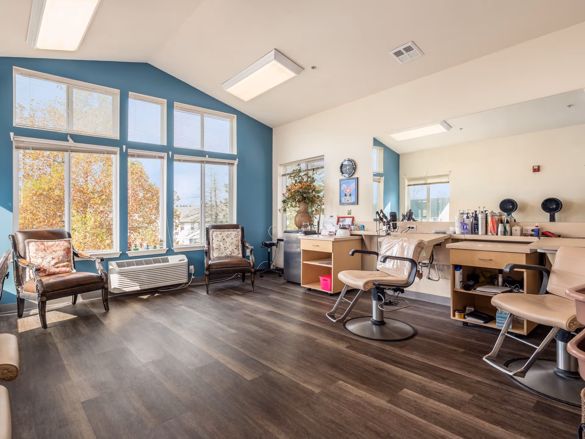 A bright and spacious salon area with large windows letting in natural light. The room features two leather chairs with floral cushions near the windows, a wall painted blue behind the windows, and salon stations with mirrors, chairs, and various hair care products on the right side. The floor is dark wood, and there is a clock and framed artwork on the wall.