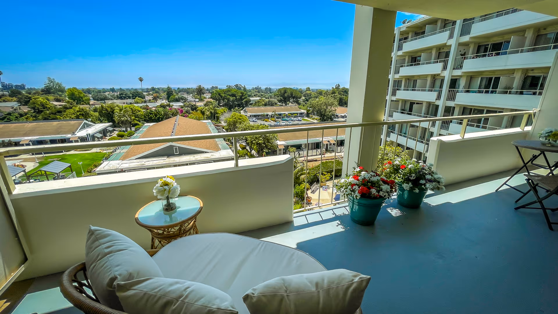 Balcony seating area with a cushioned round chair, small side table with flowers, potted blooms, and a view over the facility grounds and neighboring building.
