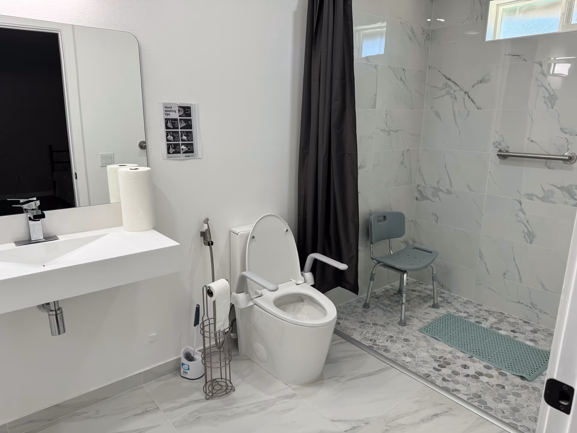 A modern bathroom with a white toilet equipped with armrests, a white sink with a faucet, a large mirror, and a roll of paper towels on the sink. There is a metal toilet paper holder with a brush next to the toilet. The shower area has a gray shower chair, a grab bar on the wall, a dark shower curtain, and marble-patterned tiles on the walls and floor.