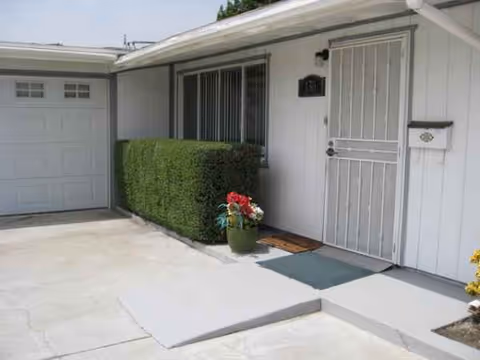 Front entrance of a light-colored senior living facility with a metal door, hedge, and potted flowers.