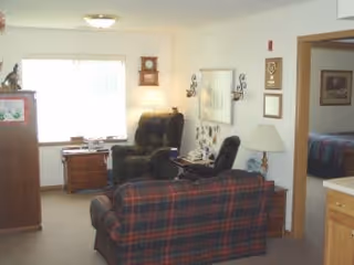 A cozy living room in an assisted living facility featuring a plaid sofa, a dark recliner chair, a wooden side table with a lamp, a wall clock, framed pictures on the walls, and a window letting in natural light. An adjacent room with a bed is partially visible through an open doorway.