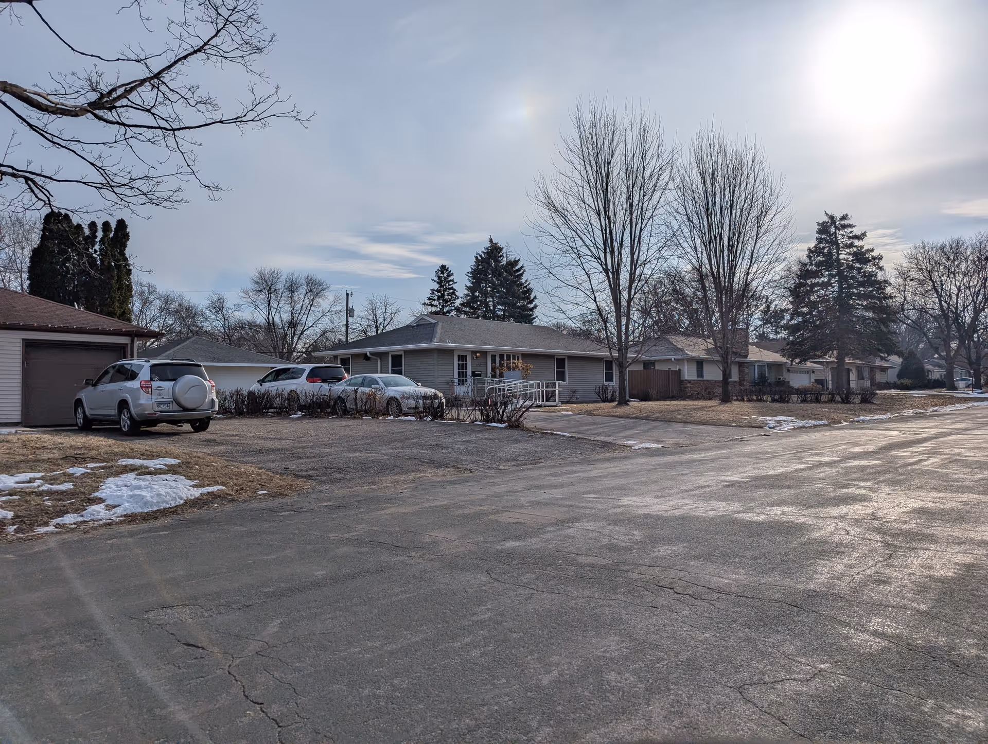 Exterior view of a single-story residential building with a ramp leading to the entrance, several parked cars, leafless trees, and patches of snow on the ground under a partly cloudy sky with the sun shining.