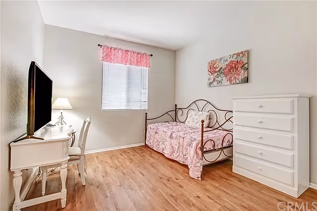 Small bedroom with a metal daybed covered in pink floral bedding, a white dresser, and a desk with a TV by a window with a pink valance.