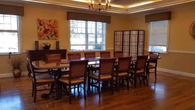 A spacious dining room with a long wooden table surrounded by ten chairs. The room has hardwood floors, three windows with blinds and valances, a chandelier hanging from the ceiling, a decorative folding screen in the corner, a sideboard with a floral arrangement, and a painting of flowers on the wall.