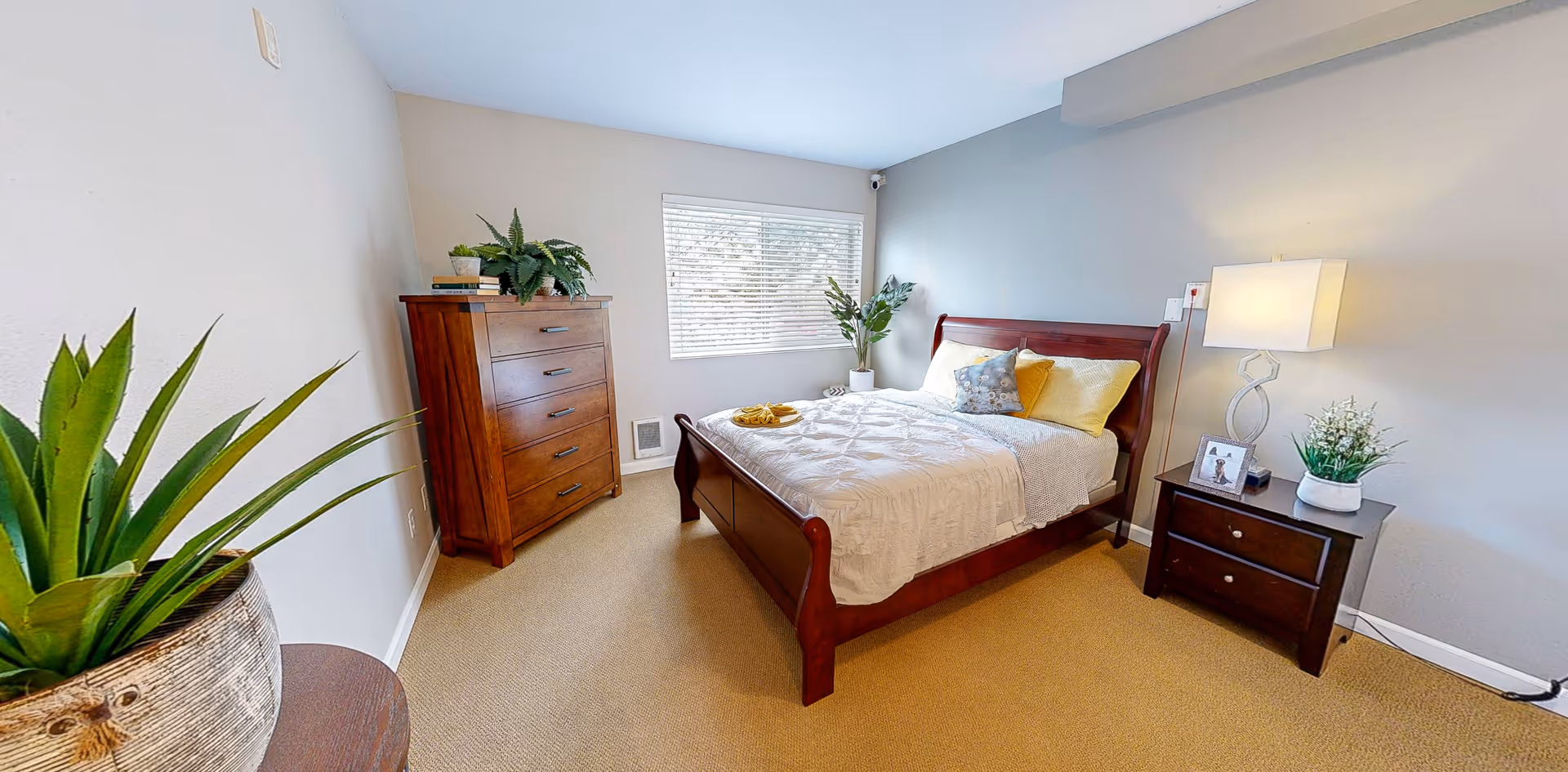Neatly furnished bedroom with a wooden bed, matching dresser and nightstand, potted plants, and a window.