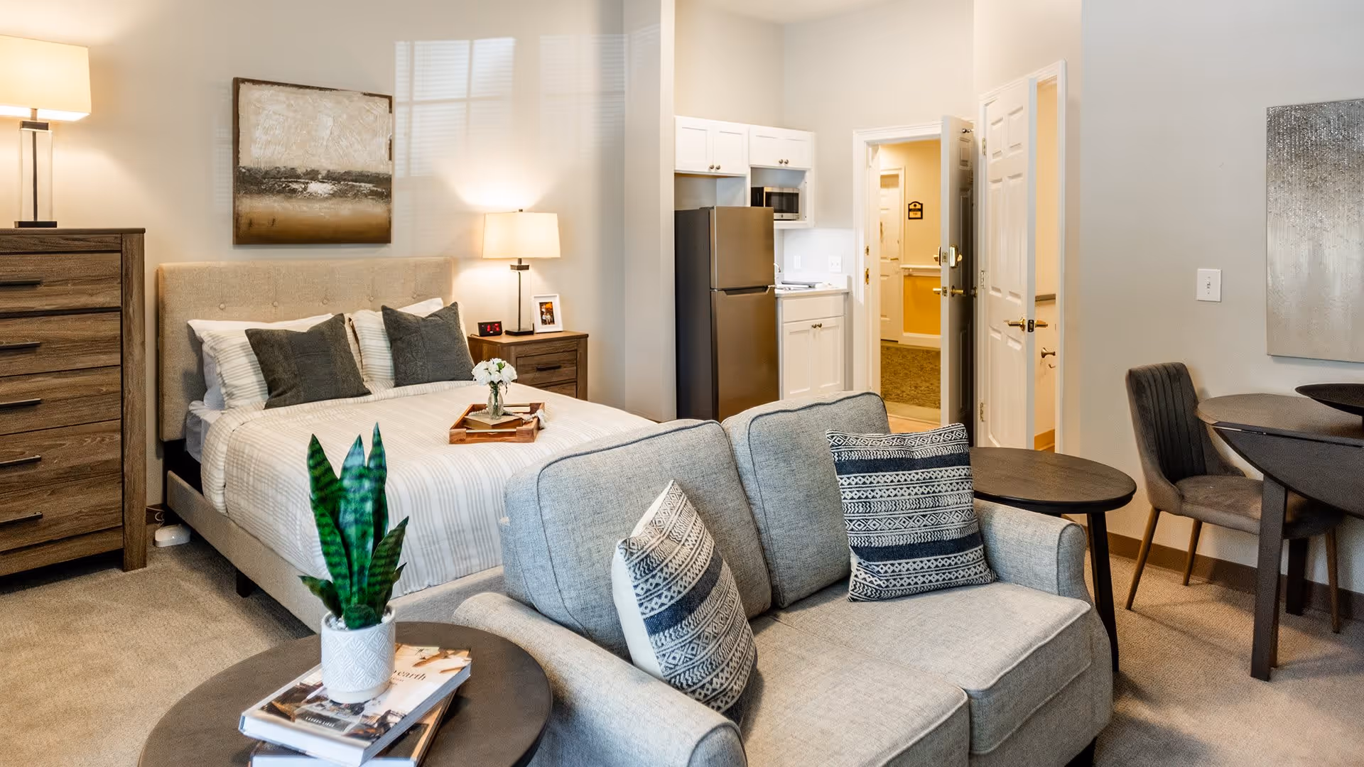 A cozy studio apartment featuring a bed with pillows and a tray with flowers, a gray sofa with patterned cushions, a wooden dresser with a lamp, a small round table with a plant and books, a kitchenette with a refrigerator and microwave, and a dining area with a round table and chairs. The room is warmly lit and decorated with wall art.