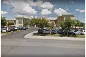 Front exterior of a large veterans center building with a parking lot, parked cars, and trees under a partly cloudy sky.