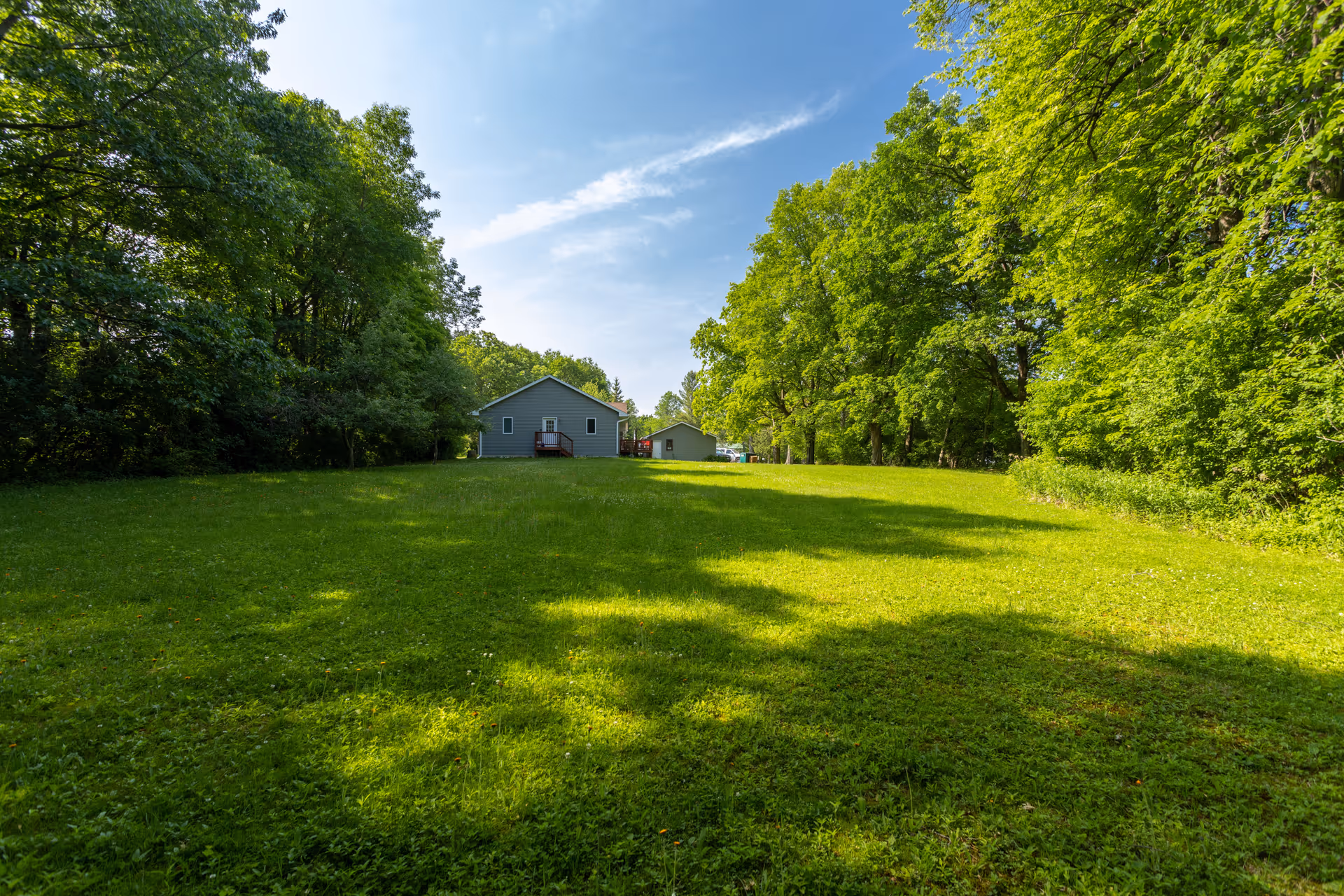 A large grassy lawn surrounded by tall green trees under a blue sky with some clouds. In the background, there are two small gray buildings with red steps leading to their entrances.