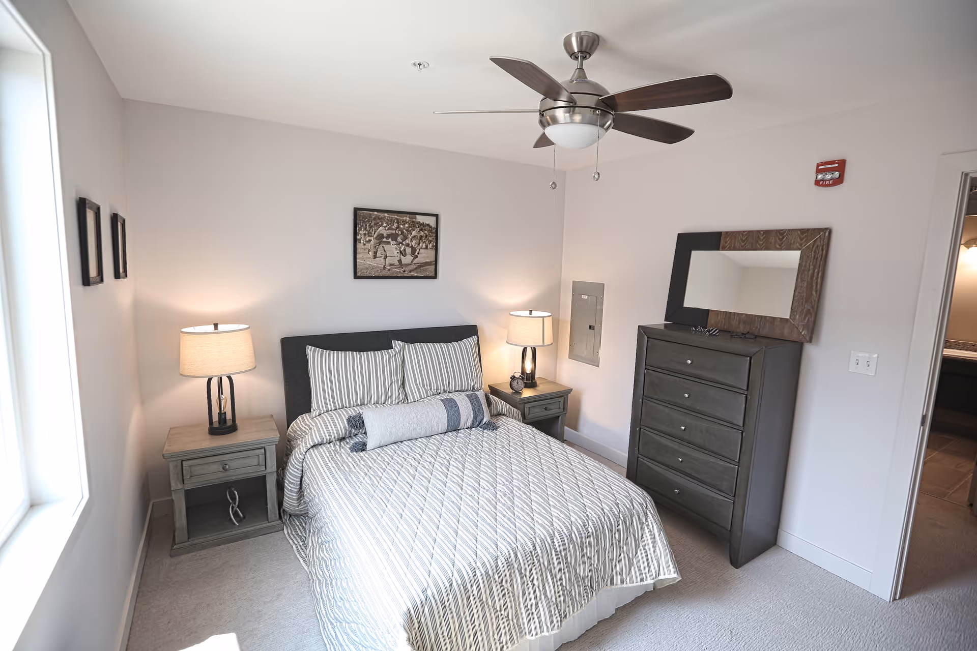 A neatly arranged bedroom with a bed covered in a striped quilt and pillows, two bedside tables each with a lamp, a dresser with a mirror on top, a ceiling fan with lights, and a framed picture on the wall above the bed. The room has light-colored walls and carpeted floor, with a window letting in natural light.
