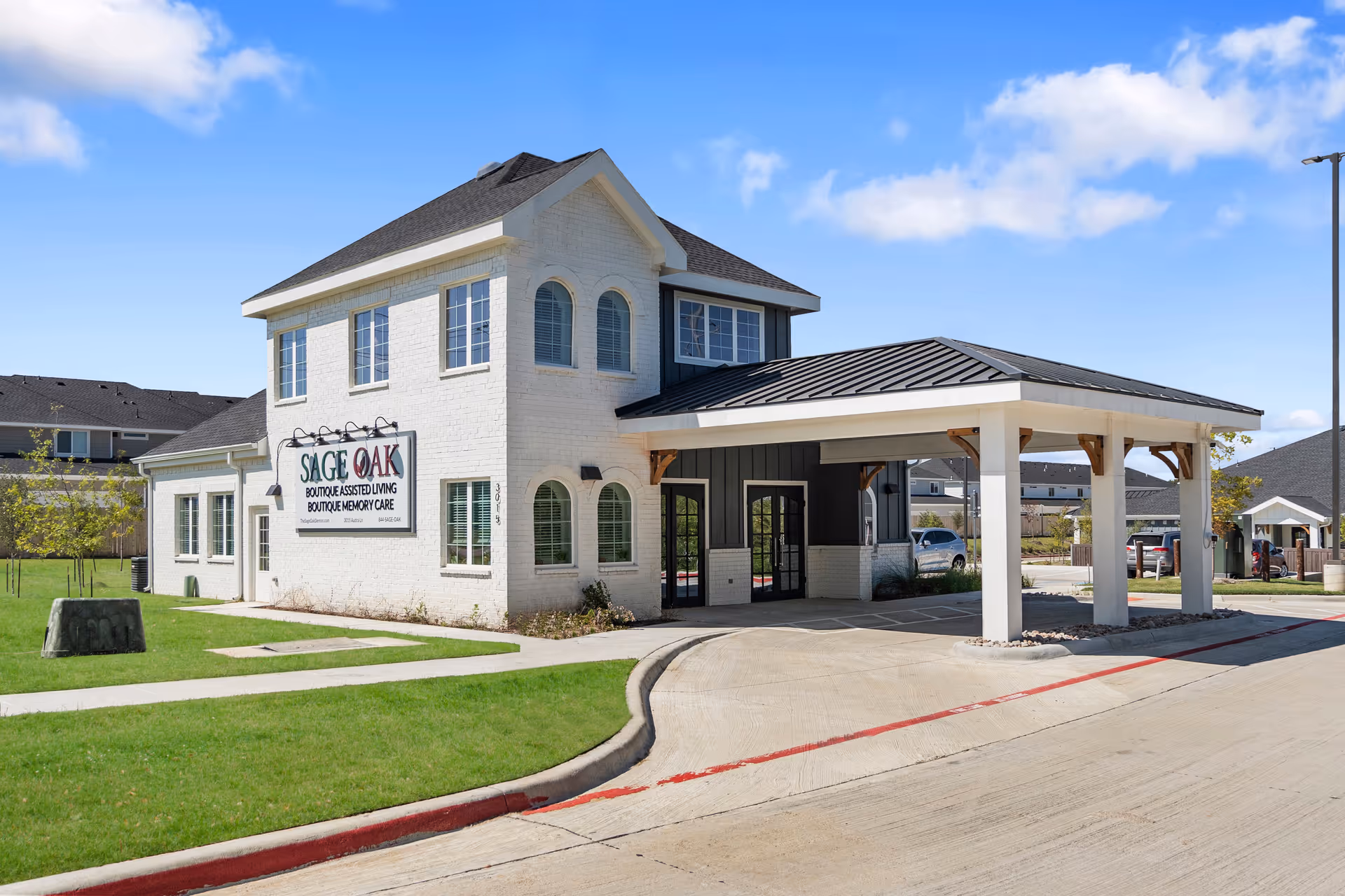 Exterior view of Sage Oak of Denton, a two-story building with white brick and dark roof. The building has a covered entrance driveway and a sign that reads 'Sage Oak Boutique Assisted Living Boutique Memory Care'. The surrounding area includes a well-maintained lawn, sidewalks, and a clear blue sky with some clouds.