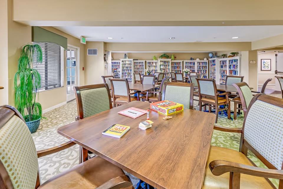 A bright and spacious common area in a senior living facility with multiple wooden tables and chairs arranged for group activities. The room features a large bookshelf filled with books along the back wall, a potted plant near a window with blinds, and a board game called 'Fact or Crap' on the table in the foreground.