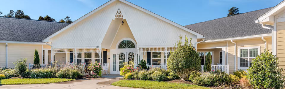 Front entrance of a light-colored senior living building with a covered porch and landscaped gardens.