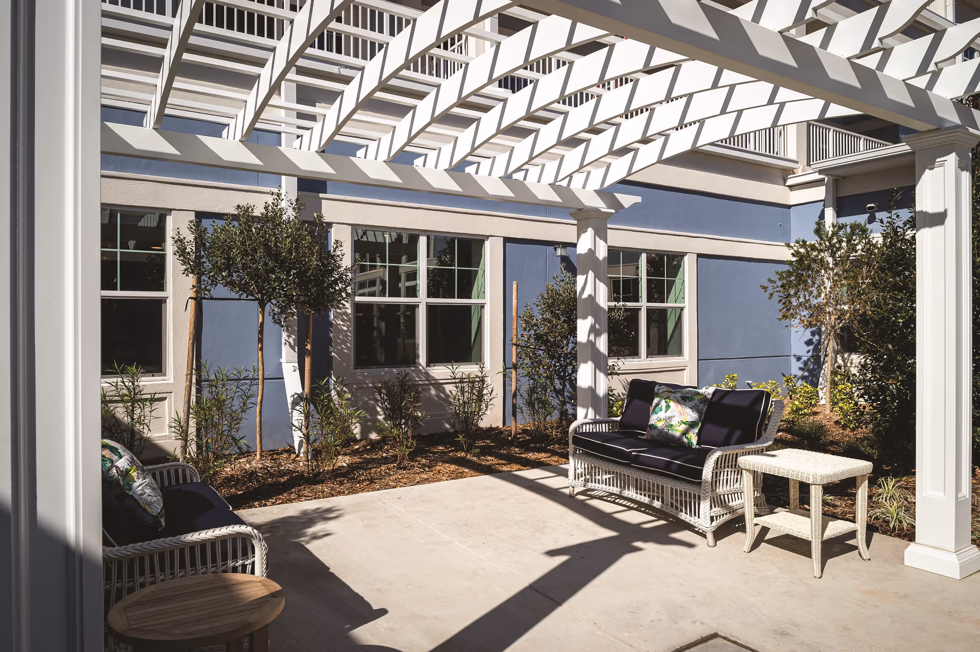 Outdoor patio area with white pergola casting shadows on the concrete floor, two white wicker sofas with dark cushions and decorative pillows, a small white wicker side table, and some small trees and plants along the building wall painted in blue and beige.