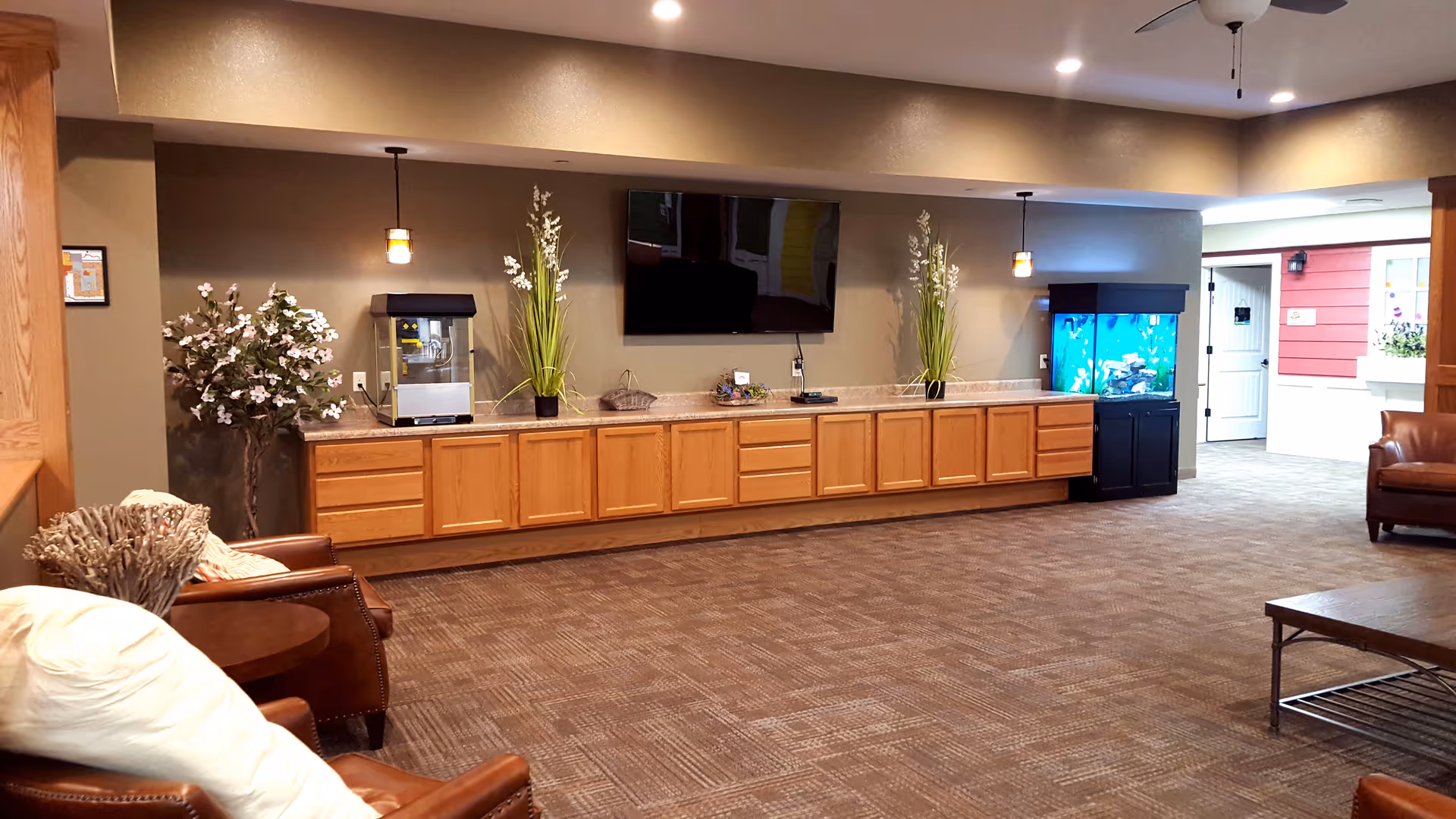 A spacious common area with brown carpet and beige walls featuring a long wooden cabinet with a countertop. On the countertop are two tall green plants, a popcorn machine, and a large fish tank. Above the cabinet is a mounted flat-screen TV. The room has leather armchairs with pillows and a wooden coffee table. There is a ceiling fan and recessed lighting, and an open doorway leads to another room with red walls and white doors.