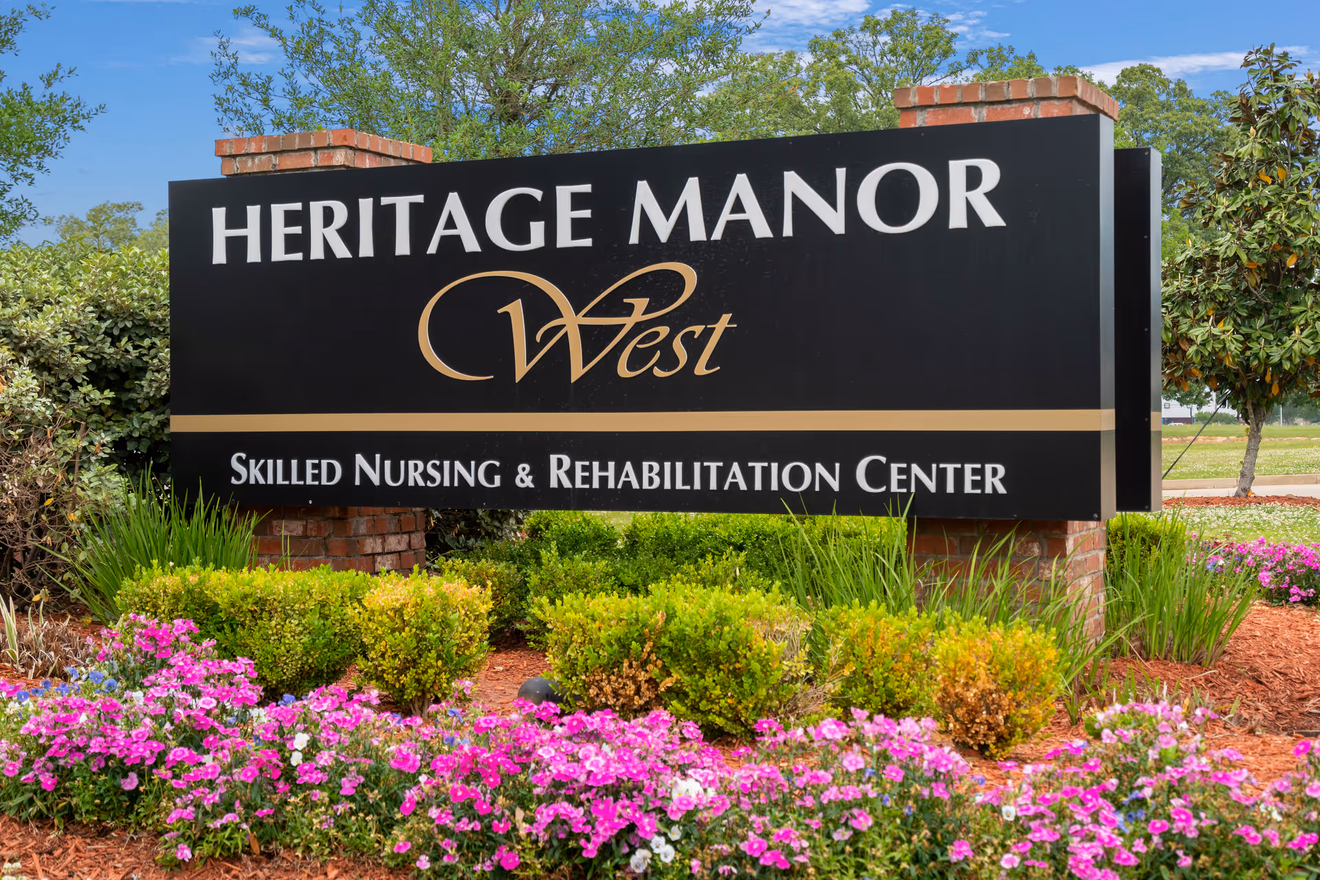 A large black sign with white and gold lettering that reads 'HERITAGE MANOR West Skilled Nursing & Rehabilitation Center' surrounded by green bushes and pink flowers with a clear blue sky in the background.