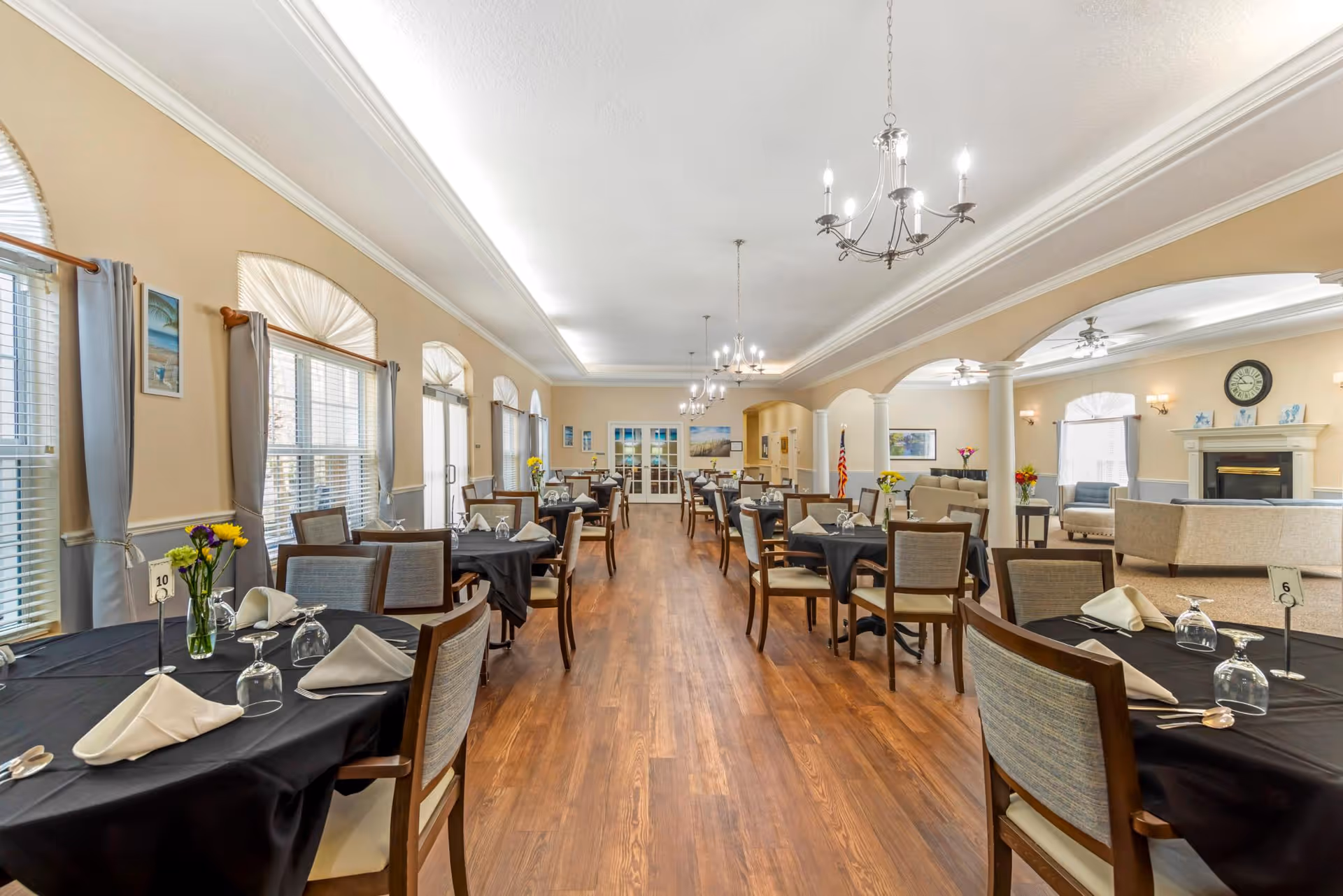 A spacious dining room with multiple tables covered in black tablecloths, each set with napkins, glasses, and silverware. The room features wooden flooring, large windows with gray curtains, chandeliers hanging from the ceiling, and a seating area with sofas and a fireplace in the background.