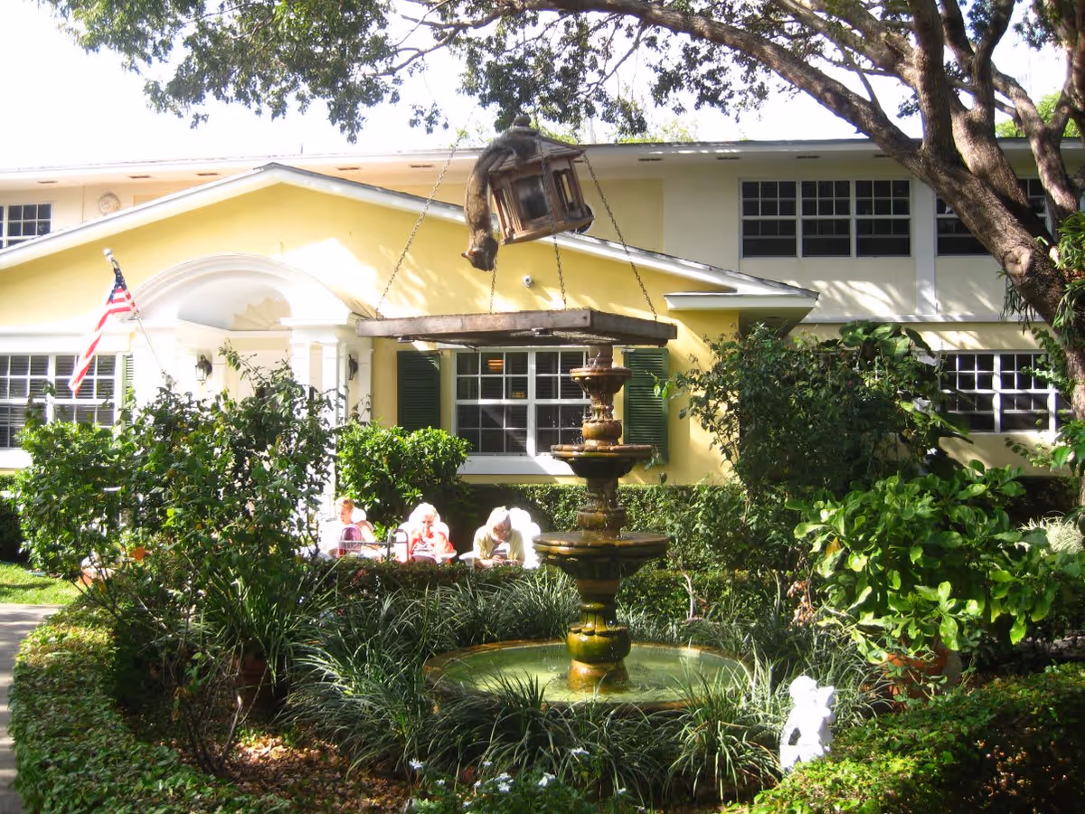 A yellow two-story building with white trim and green shutters surrounded by lush greenery and trees. In front of the building is a tiered water fountain with a hanging lantern above it. Three elderly people are seated on a bench near the building, partially obscured by bushes. An American flag is displayed near the entrance.