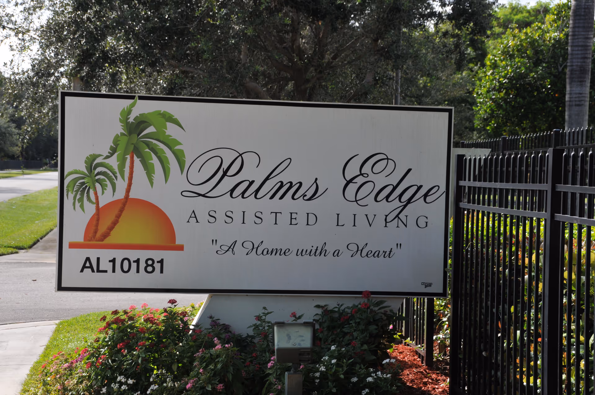 Outdoor sign for Palms Edge Assisted Living featuring a graphic of two palm trees and a setting sun, with the tagline 'A Home with a Heart'. The sign is surrounded by greenery and flowers, with a black metal fence to the right.