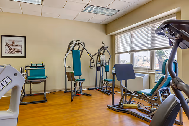 A fitness room with various exercise machines including a multi-station weight machine, a leg press, and other strength training equipment. The room has wooden flooring, beige walls, a large window with blinds letting in natural light, and a framed picture on the wall.