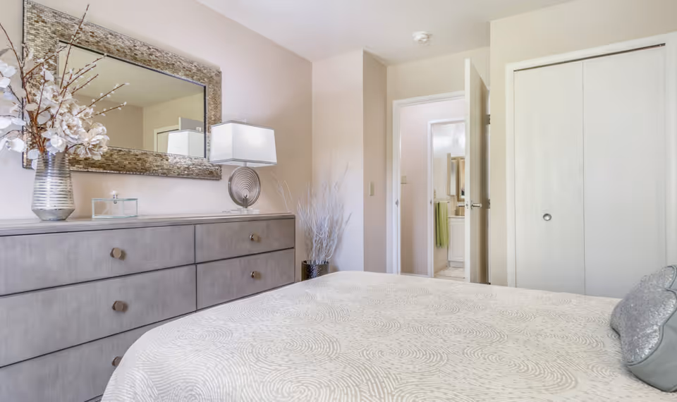 A bright bedroom with a large bed covered in a patterned white bedspread and a decorative pillow. To the left is a gray dresser with six drawers, topped with a vase of white flowers, a glass box, and a modern lamp. Above the dresser hangs a large rectangular mirror with a textured frame. In the background, there is an open door leading to a bathroom with a green towel visible, and a closed white closet door.