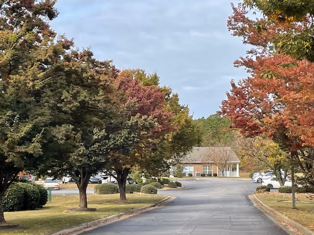 A paved driveway lined with trees showing autumn foliage in shades of green, red, and yellow. Several parked cars are visible along the sides of the driveway. In the background, there is a single-story brick building with white trim and a porch.
