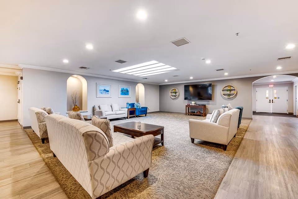 Spacious senior living facility common area with multiple beige and patterned sofas arranged around a wooden coffee table on a large carpet. The room has light-colored walls, recessed lighting, a skylight, and a flat-screen TV mounted on the far wall with decorative shelves on either side. There are framed pictures on the wall and a hallway leading to double doors in the background.